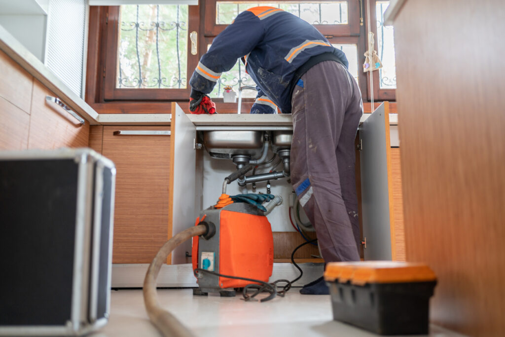 A plumber wearing a blue jacket is working under a kitchen sink in Delavan, WI, with a utility box and a vacuum machine connected to the pipes nearby for effective drain cleaning.